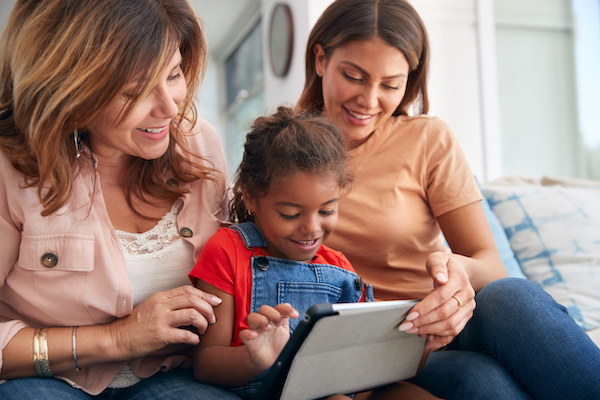 Multi-Generation Female Hispanic Family Sitting On Sofa At Home Using Digital Tablet